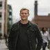 Man wearing a black jacket and jeans standing outdoors with a blurred background